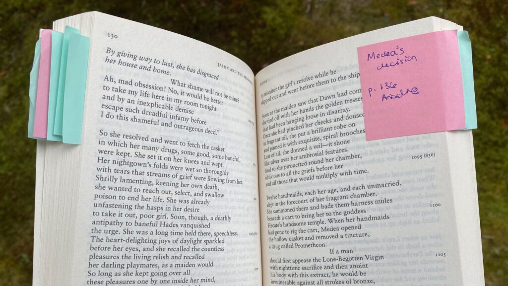 A paperback copy of Apollonius' Argonautica lies open, with a pink sticky note labeled with a page number and 'Medea's choice' stuck to the page